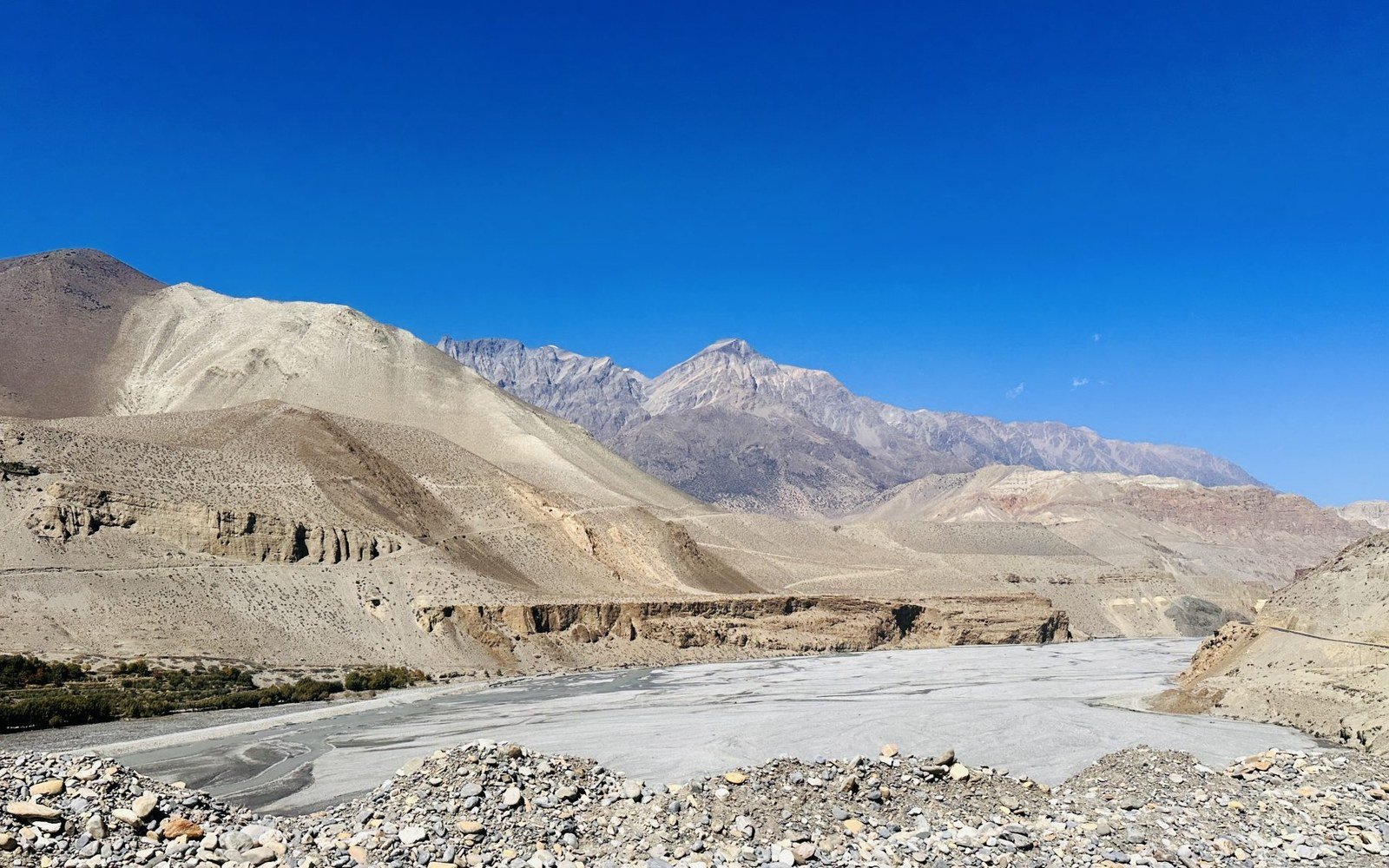 The broad braided gravel bed of a river running between steep eroded hillsides in Nepal