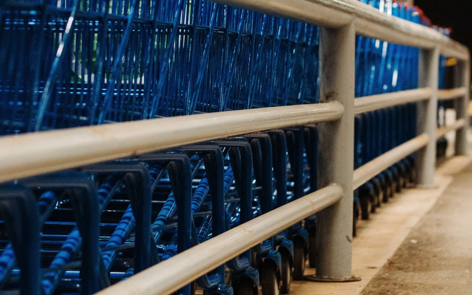 A long line of nested blue shopping trolleys standing behind a painted steel guard rail under a canopy, photographed low along the row with no lettering visible on any handle