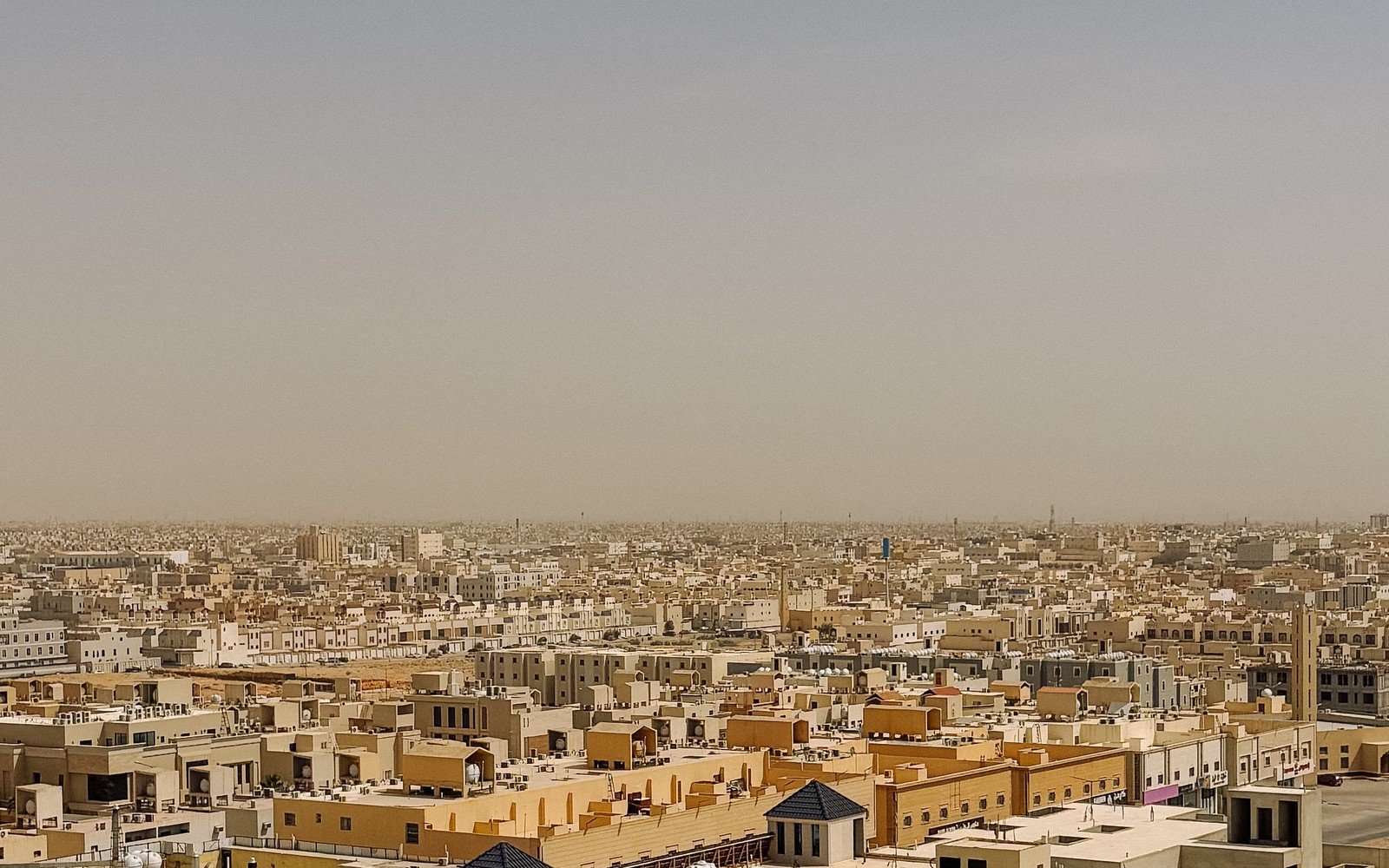 A wide view across a low rise city of sand coloured flat roofed buildings under a pale hazy sky, rooftop water tanks and air conditioning units in the foreground and the built up plain running to the horizon