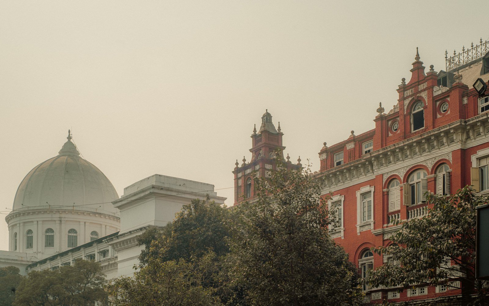 Colonial-era buildings in Kolkata under a heavy haze, a red brick corner block with a turret on the left and a domed white building behind trees
