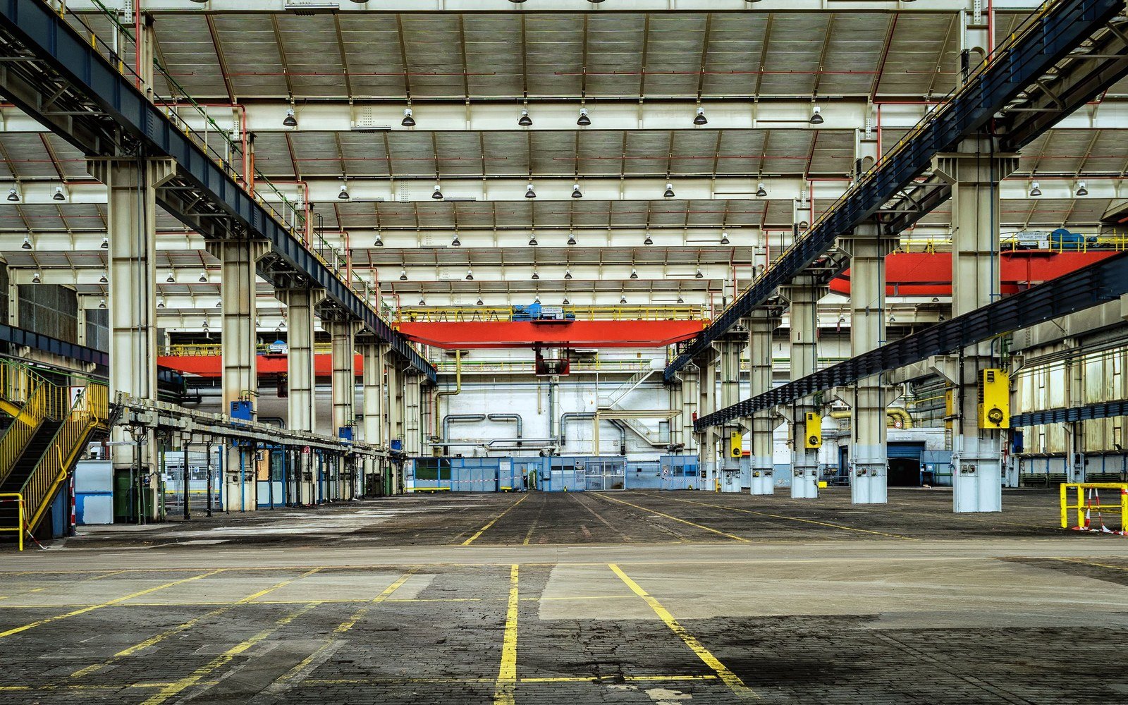 The empty floor of a large industrial hall with an overhead travelling crane, painted yellow bay markings on bare concrete and plant along both walls