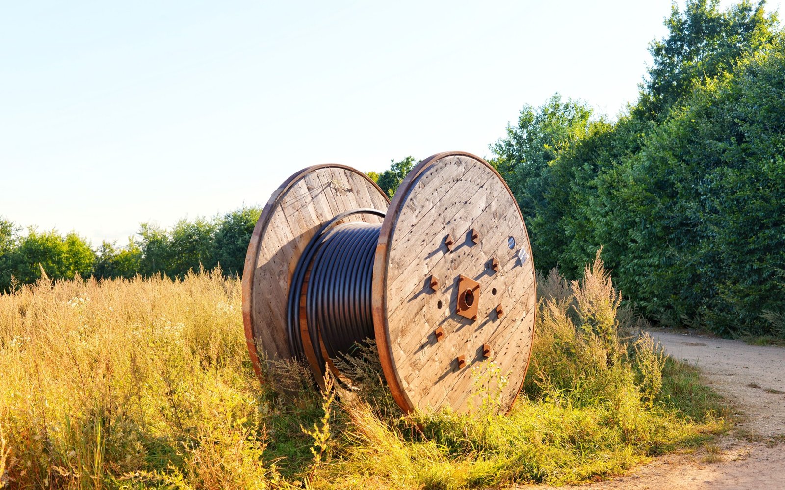 A large wooden cable reel wound with black cable, standing in rough grass beside a track on a bright summer evening