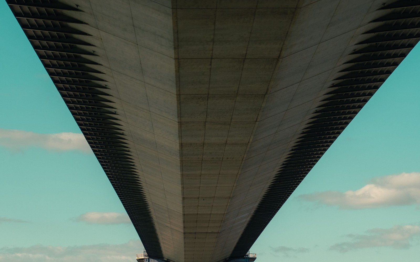 The underside of a long suspension bridge deck seen from below, narrowing towards a distant concrete tower against a pale sky