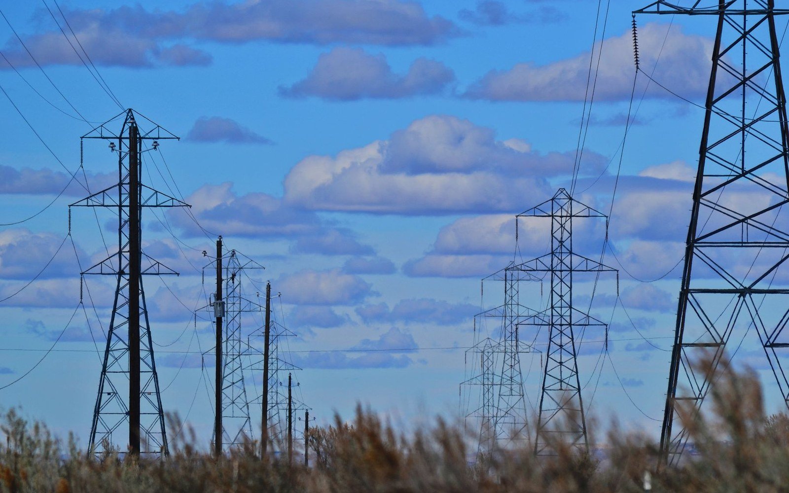 Lines of steel electricity transmission towers carrying conductors against a bright cloudy sky