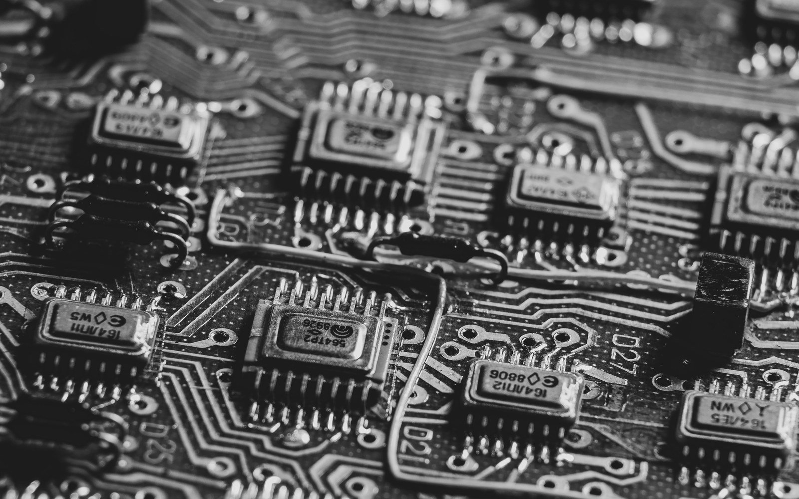 A monochrome close view across a printed circuit board, with surface mounted components, solder pads and etched copper tracks running out of focus towards the edge of frame