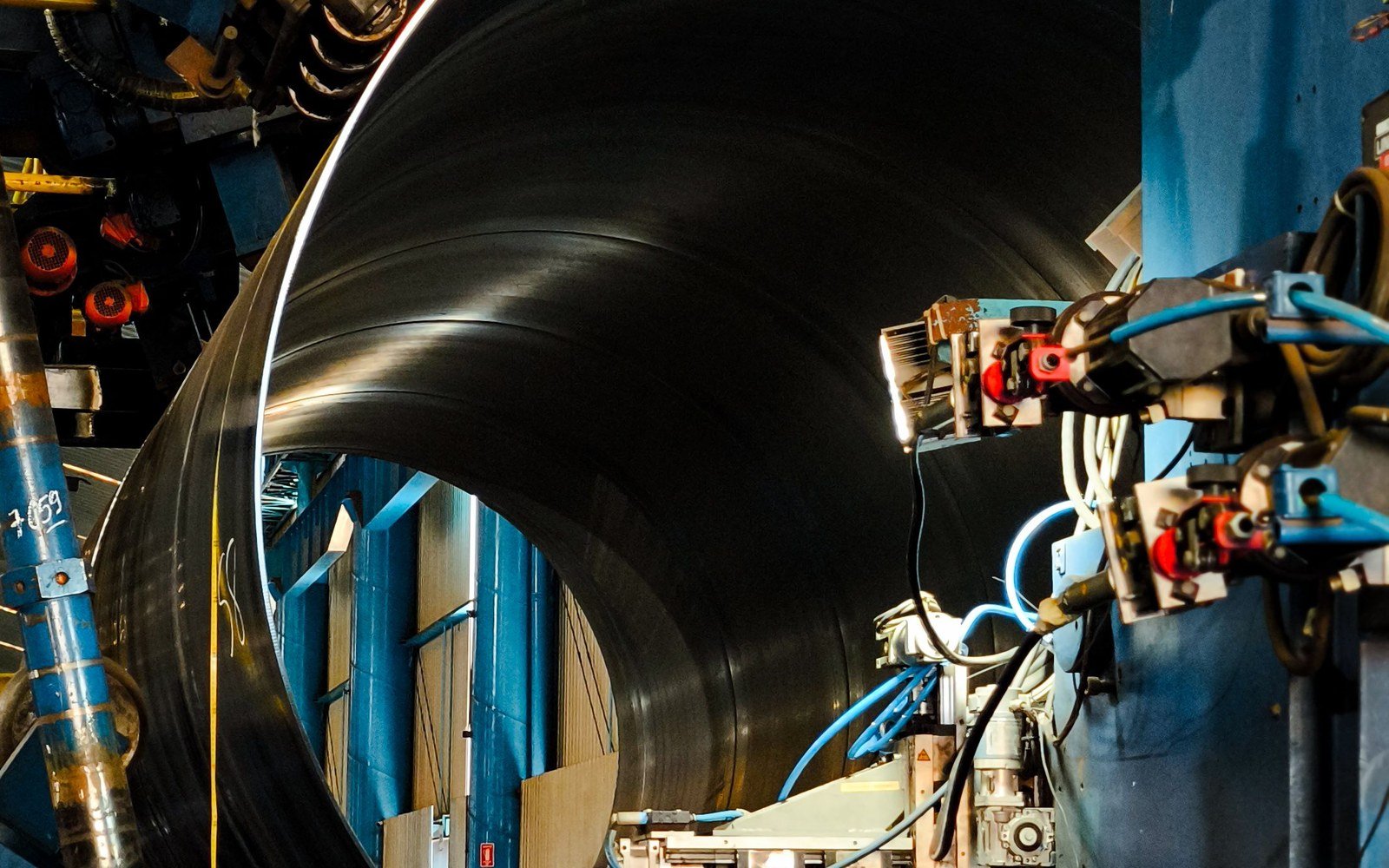 The open mouth of a large steel pipe passing through a spiral welding machine inside a factory, with blue framework, hydraulic lines and welding heads around the pipe