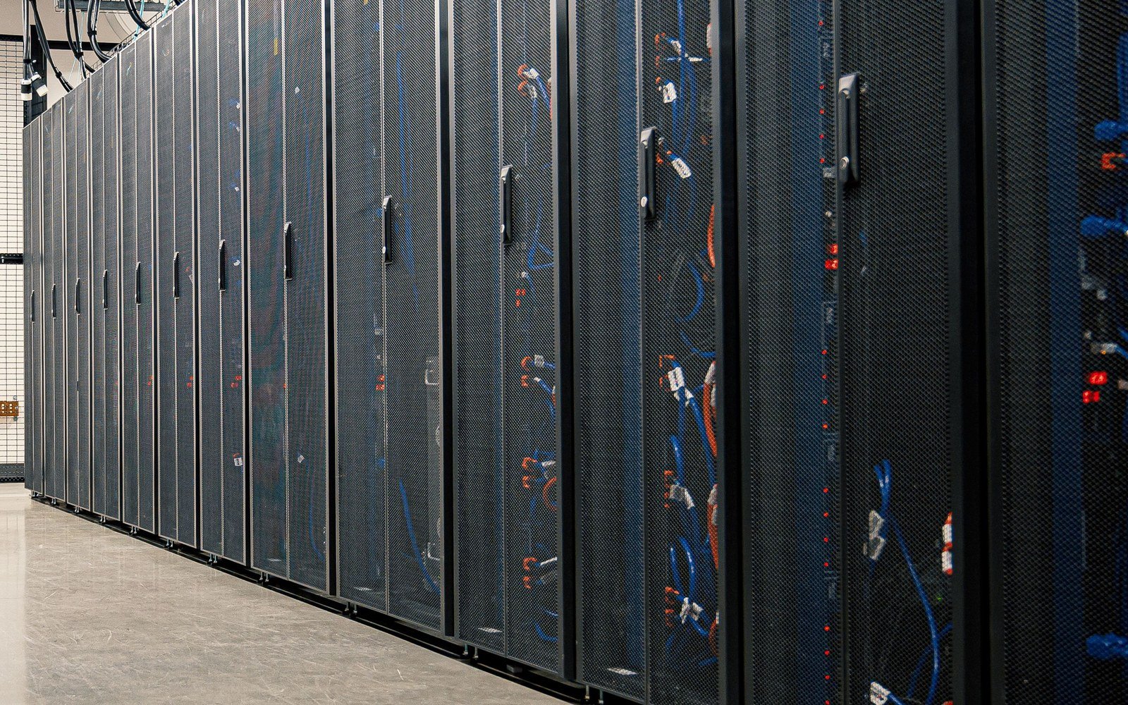 A row of black perforated server cabinet doors receding down a data hall aisle, blue and orange patch cables visible through the mesh and small red status lights along one frame