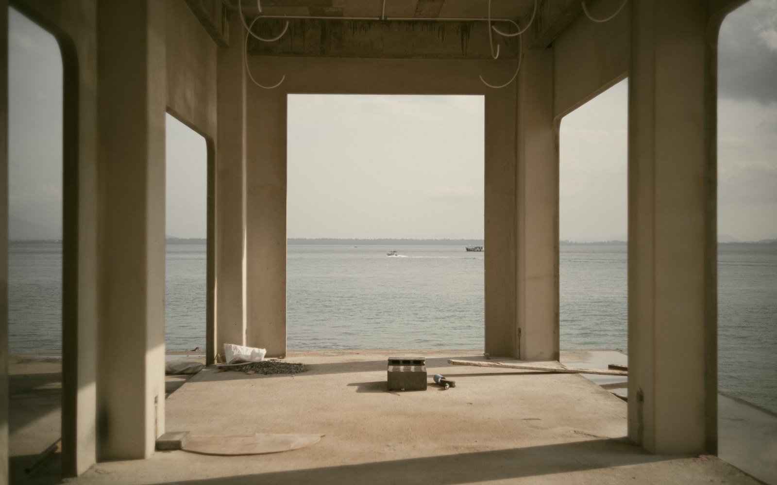 The ground floor of a reinforced concrete house frame under construction, three square columns and a flat soffit framing an open view of calm sea and a low horizon, an unfinished floor slab underfoot and formwork residue at the column bases