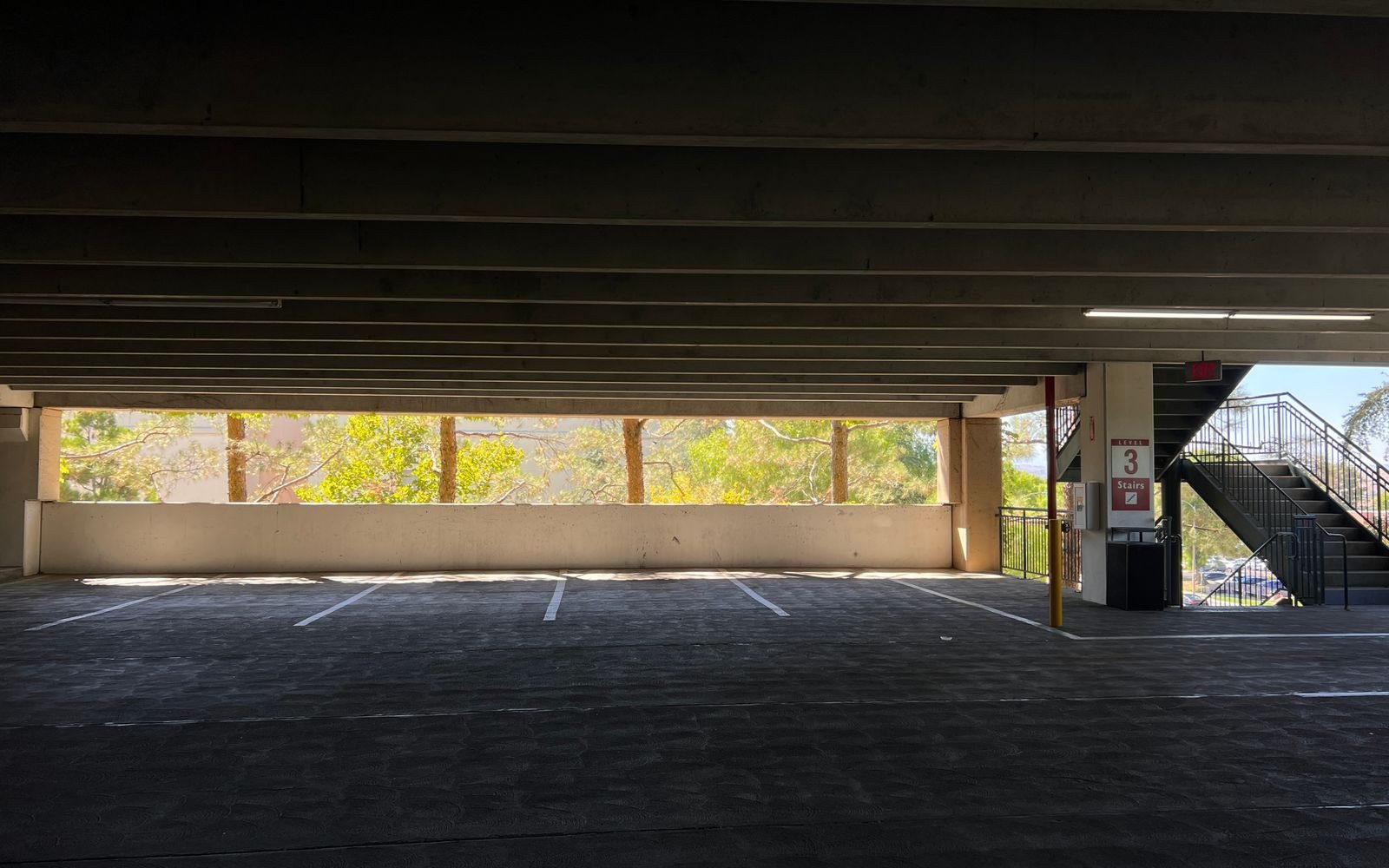 Empty level of a multistorey car park in daylight, bare concrete beams overhead and marked bays running to an open edge, with an external stair and a level number sign at the far end