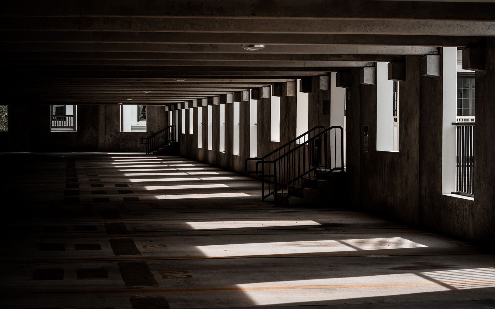 An empty upper deck of a concrete multi-storey car park, sunlight falling in bands through the open bays onto a bare deck with painted bay numbers and a steel handrail
