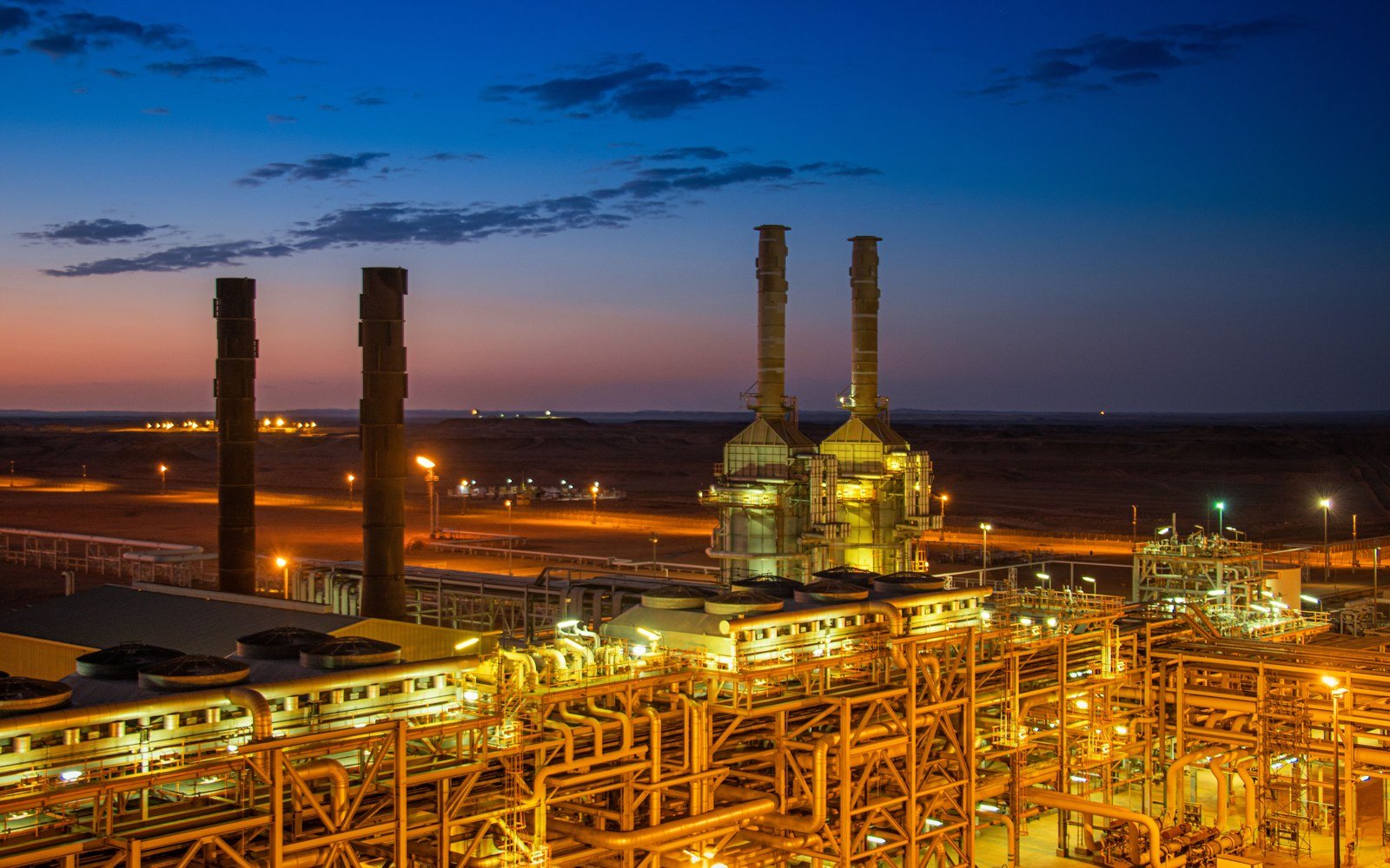 A lit gas processing plant standing on flat open ground at dusk, two tall stacks against a pink and blue sky, with banks of air cooled exchangers and pipe racks picked out in orange light below them