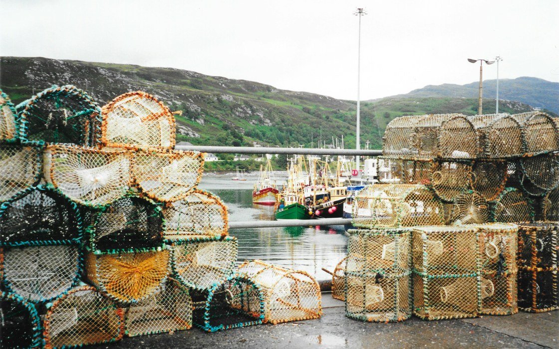 Stacked fishing creels on a wet quayside at Ullapool harbour in the Scottish Highlands, with fishing vessels moored beyond and hills behind