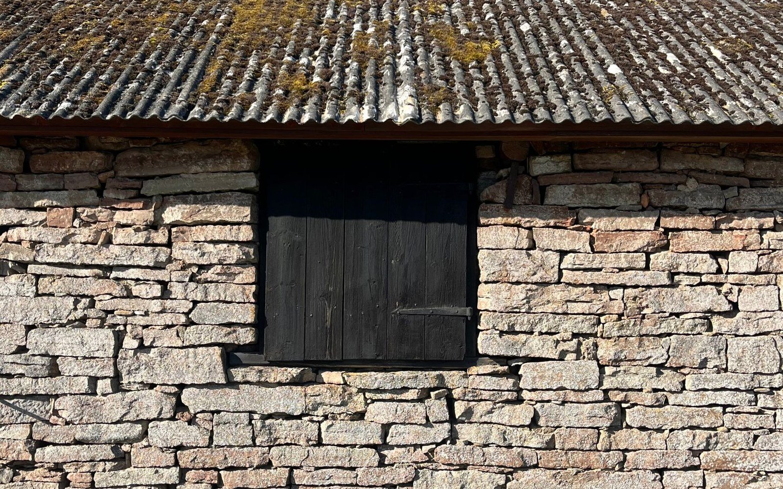 A dark shuttered opening set into a rubble stone wall beneath a mossy stone roof