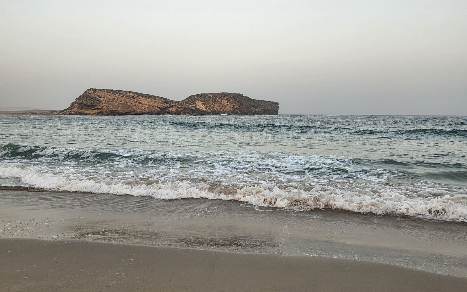 A twin rock islet off a sandy beach at Salalah in Oman, with surf breaking under a pale sky