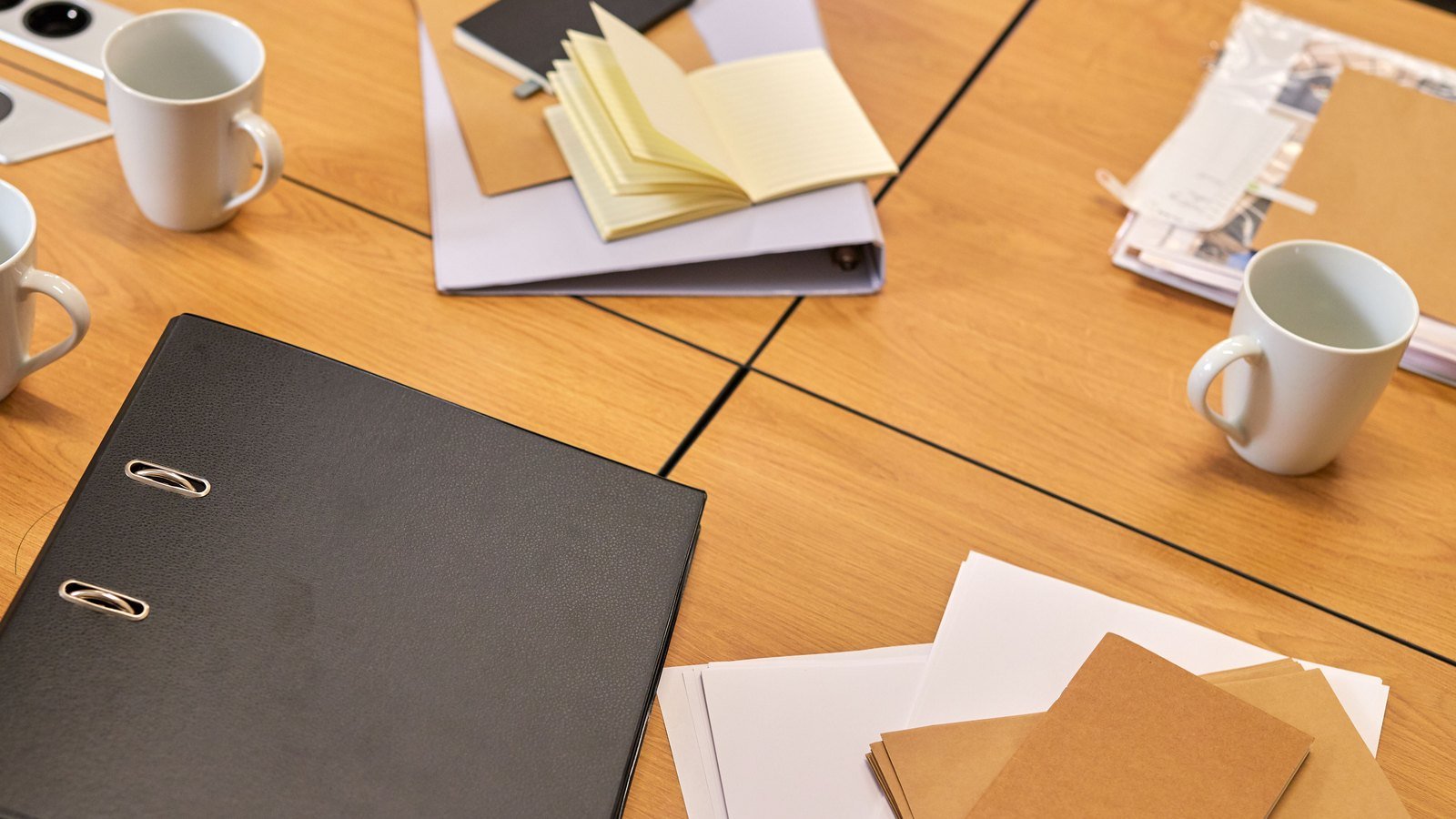 Lever arch files, blank paper, notebooks and mugs spread across a light wood meeting table