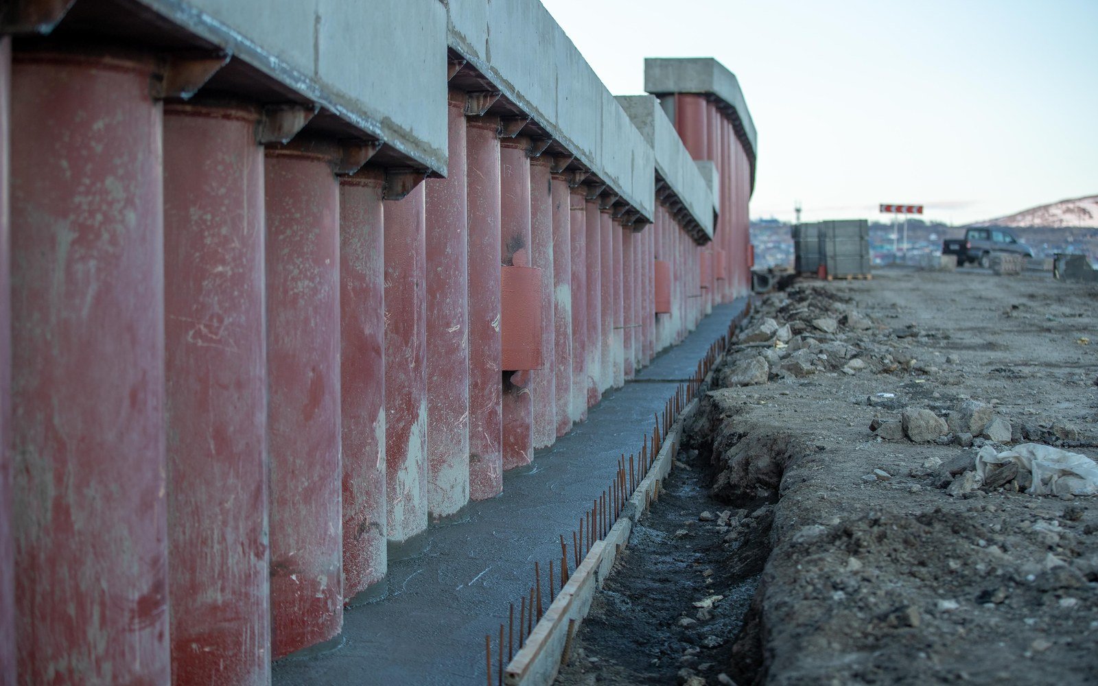 A line of concrete retaining wall panels on a cleared earthwork site, with stacked precast units and broken ground beyond