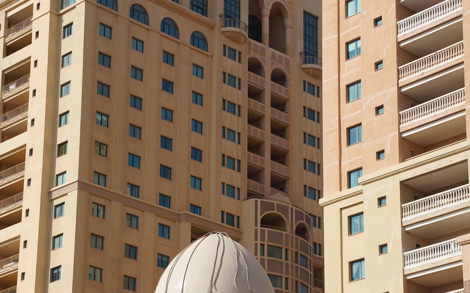 Two tall sand coloured buildings seen close up in flat daylight, regular rows of small square windows across both elevations, white balustraded balconies stepping down the right hand block, an arched loggia and a turret between them, and the white ribbed shell of a dome rising into the bottom of the frame
