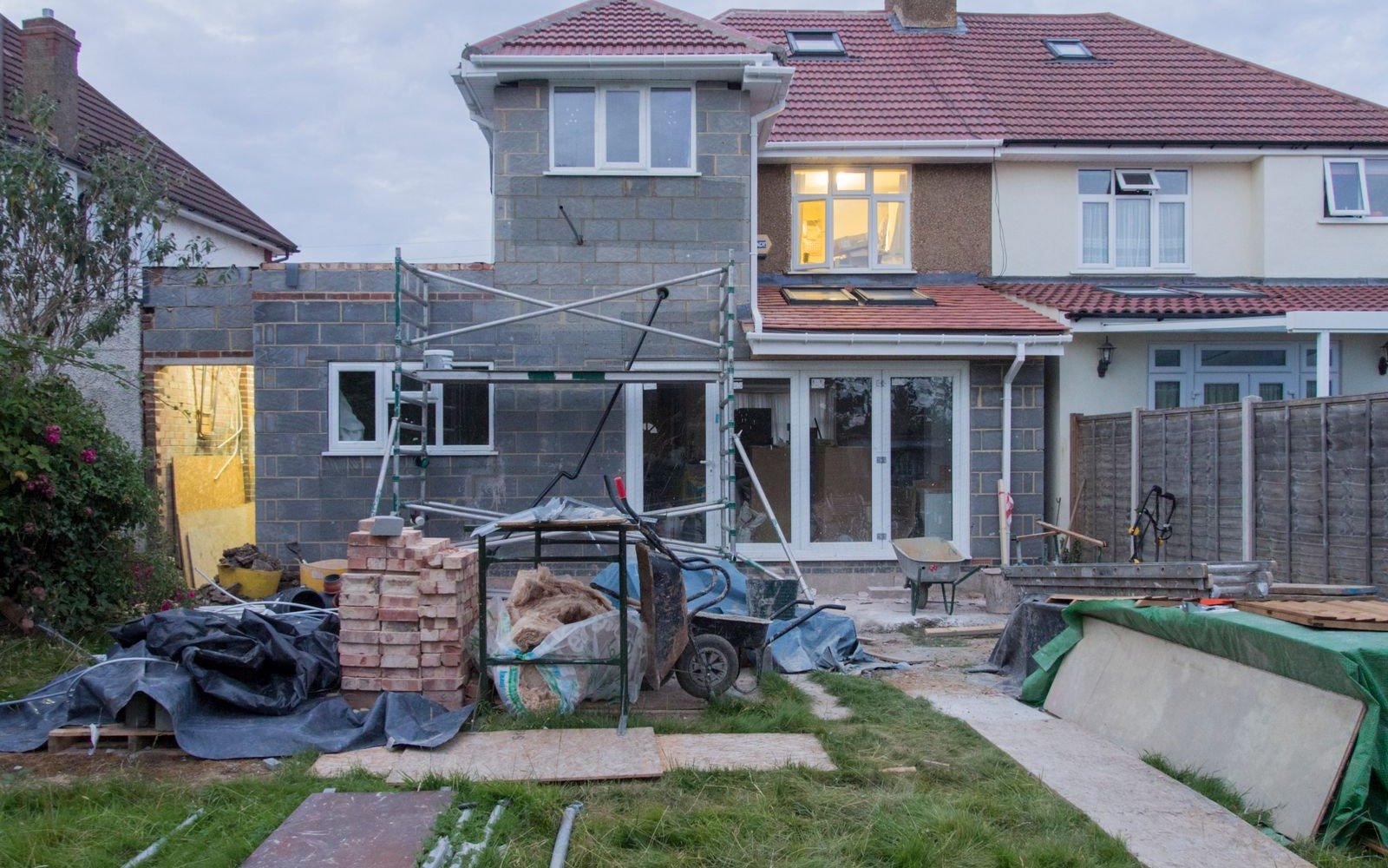 A house with a blockwork rear extension under construction, a scaffold tower and stacked bricks in the garden