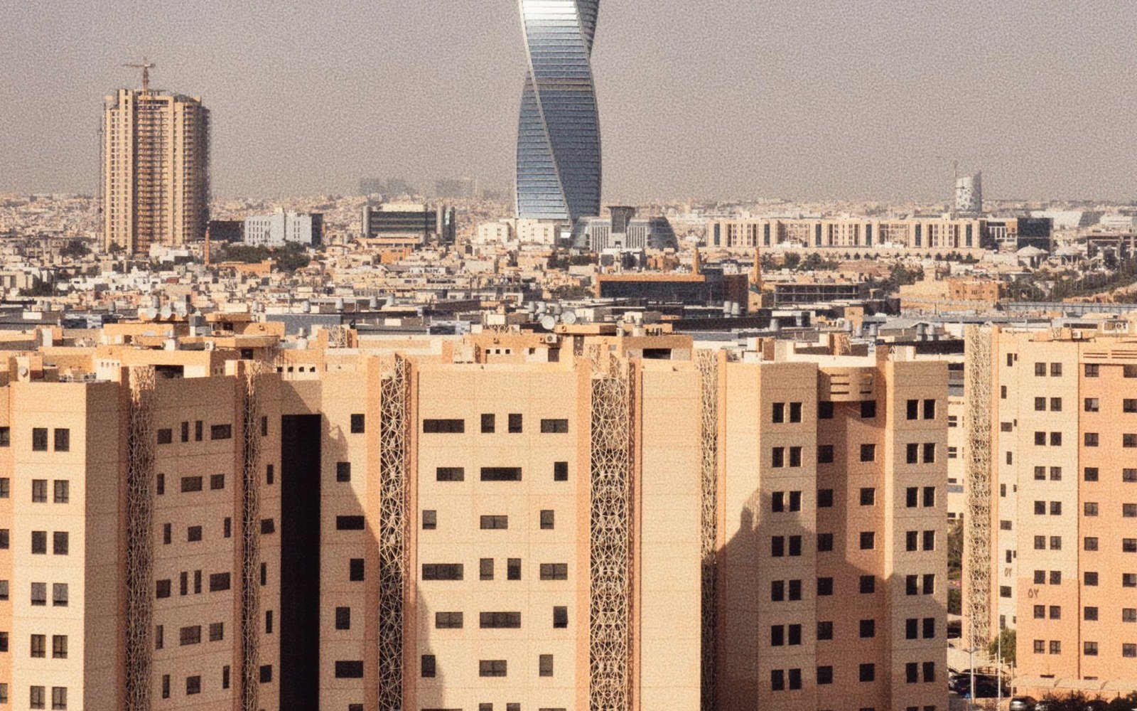 A city skyline of pale stone residential and commercial blocks with a twisting tower behind them and a tower crane on the left, seen in flat daylight haze