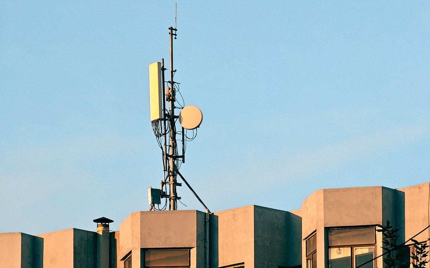 A panel antenna and a small dish on a slim mast fixed to the roof parapet of a residential block, its upper storeys lit by low evening sun