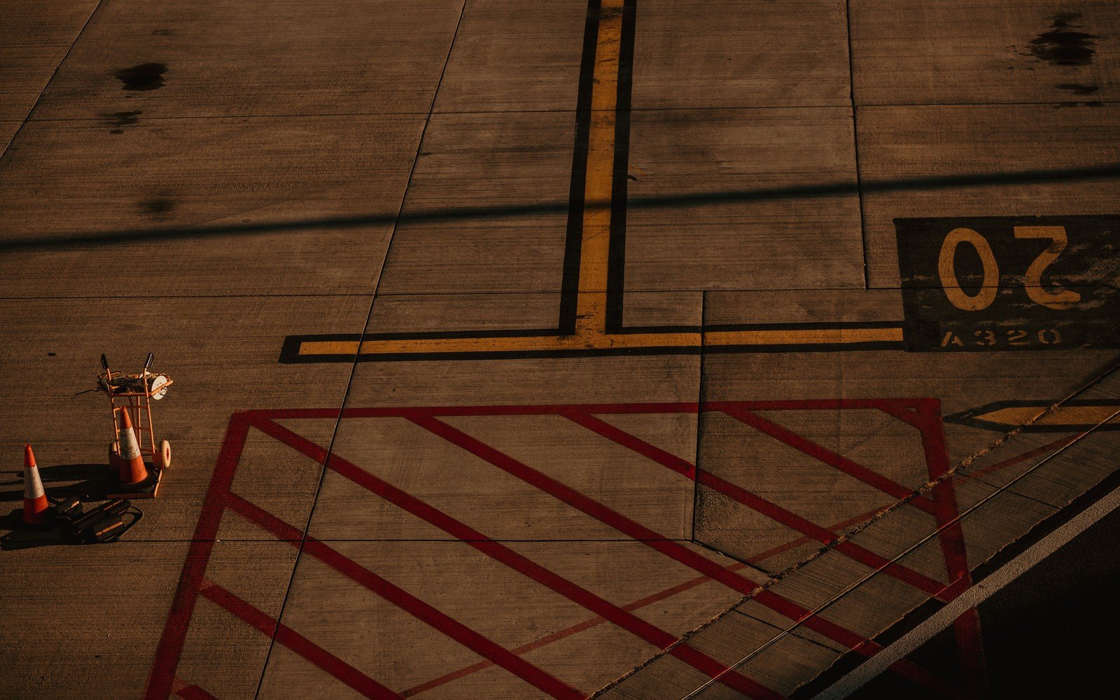 Concrete apron markings in low evening light, a yellow taxi lane crossing a red hatched stand box, the stand number and the aircraft type stencilled on the slab and two traffic cones with a hand trolley at the left edge