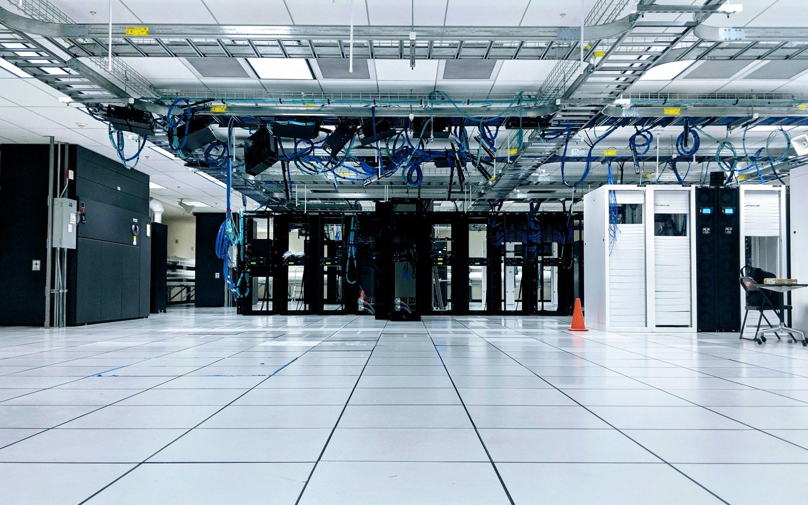 Rows of dark server cabinets standing on a raised floor in a data hall, with blue patch cabling carried on trays above the aisle and lit ceiling panels overhead