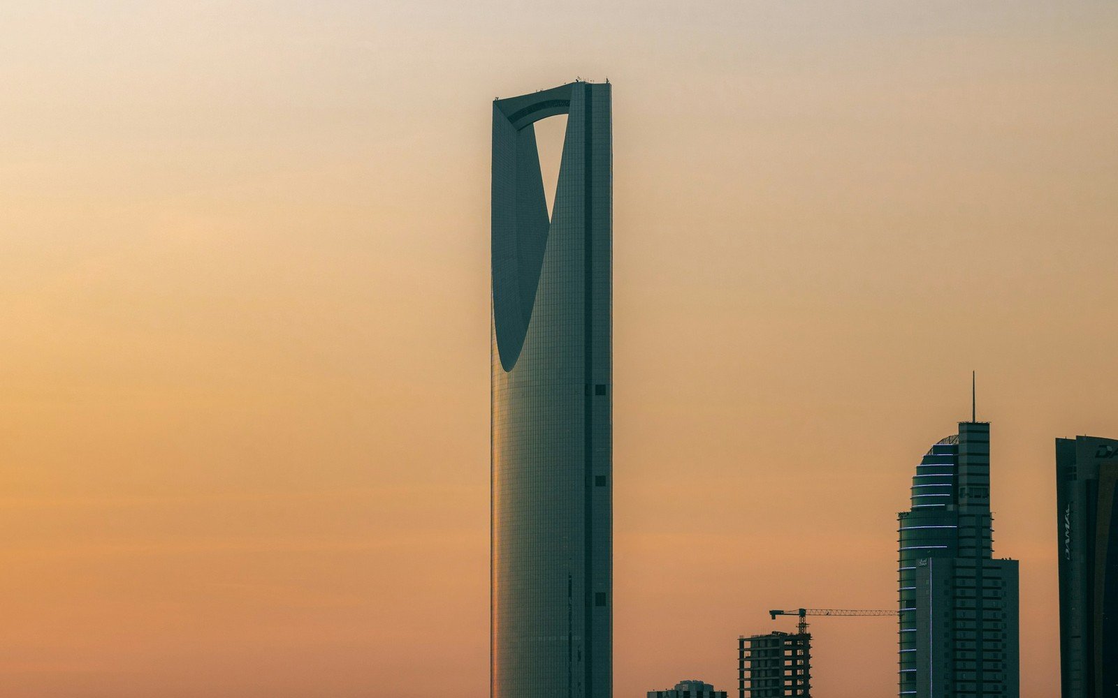 The Kingdom Centre tower in Riyadh silhouetted against an orange dusk sky, with residential towers and a tower crane on the skyline to the right
