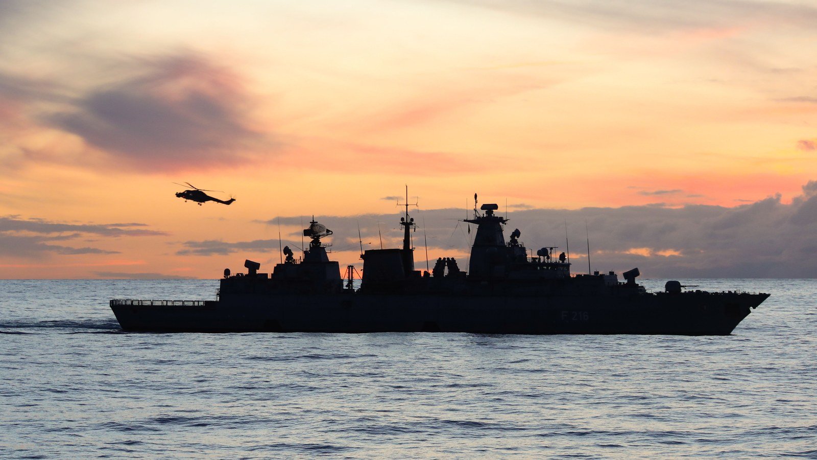 A warship silhouetted at sea at sunset with a helicopter above