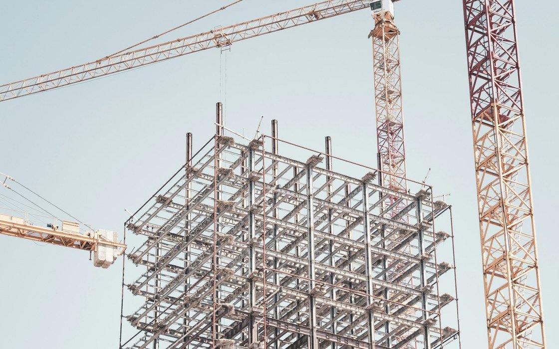 A steel and concrete building frame under construction with two tower cranes standing over it against a clear sky