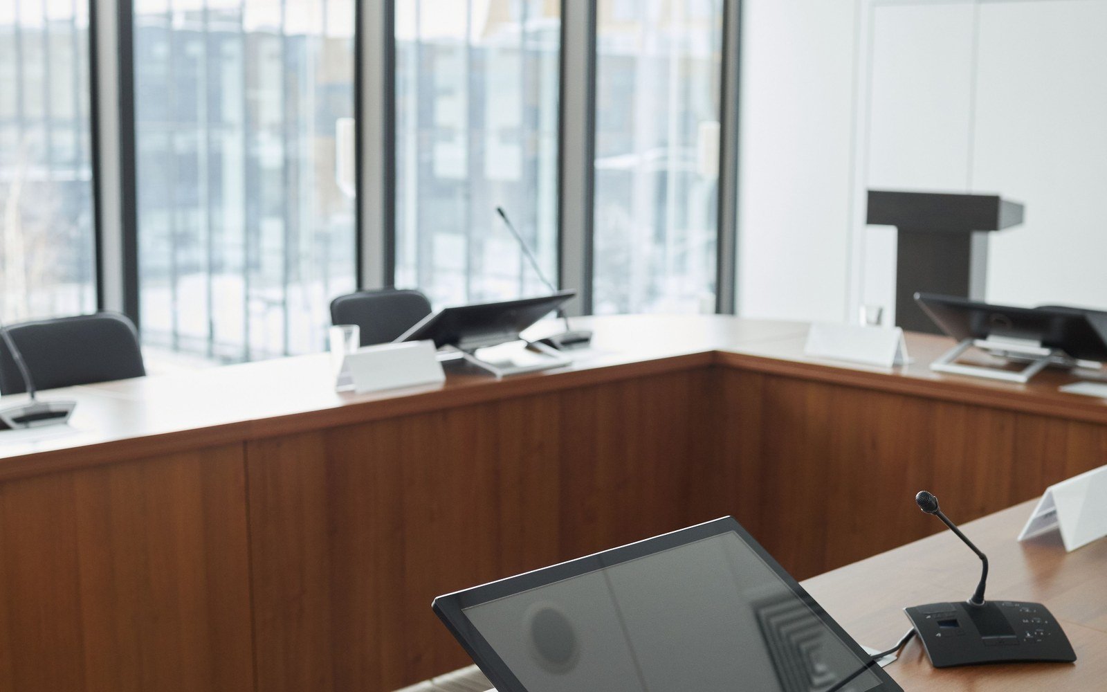 An empty modern hearing room with a curved timber table, desk microphones and screens, daylight entering from a full height window