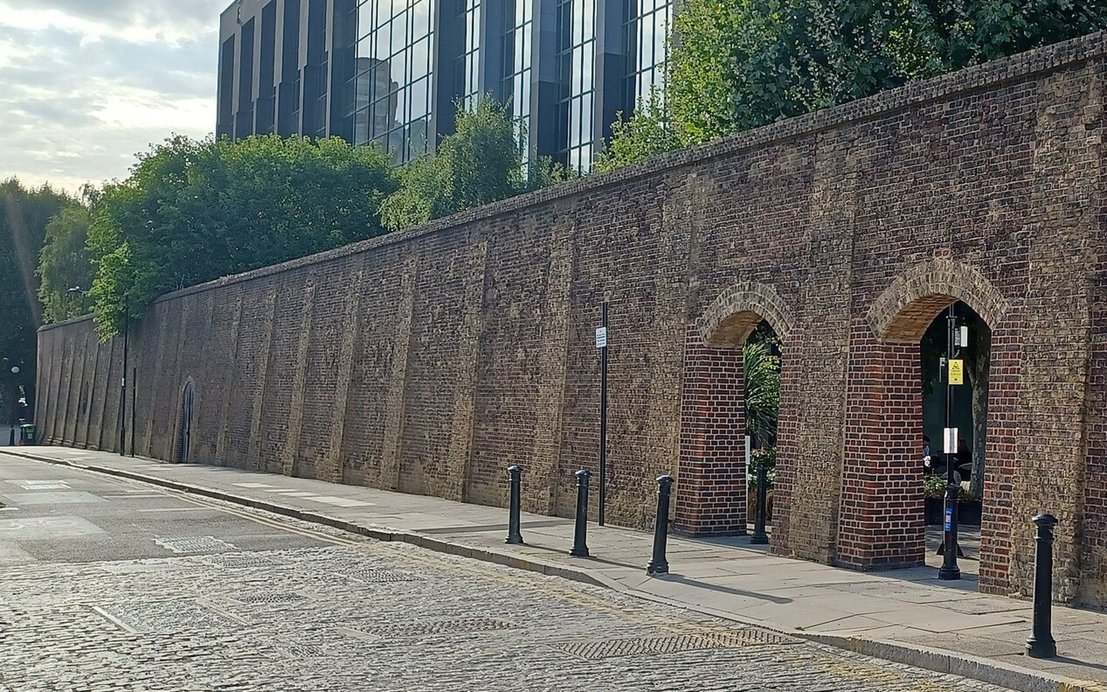 A long brick boundary wall with blind arches running beside a cobbled street, with a glazed office building rising behind it