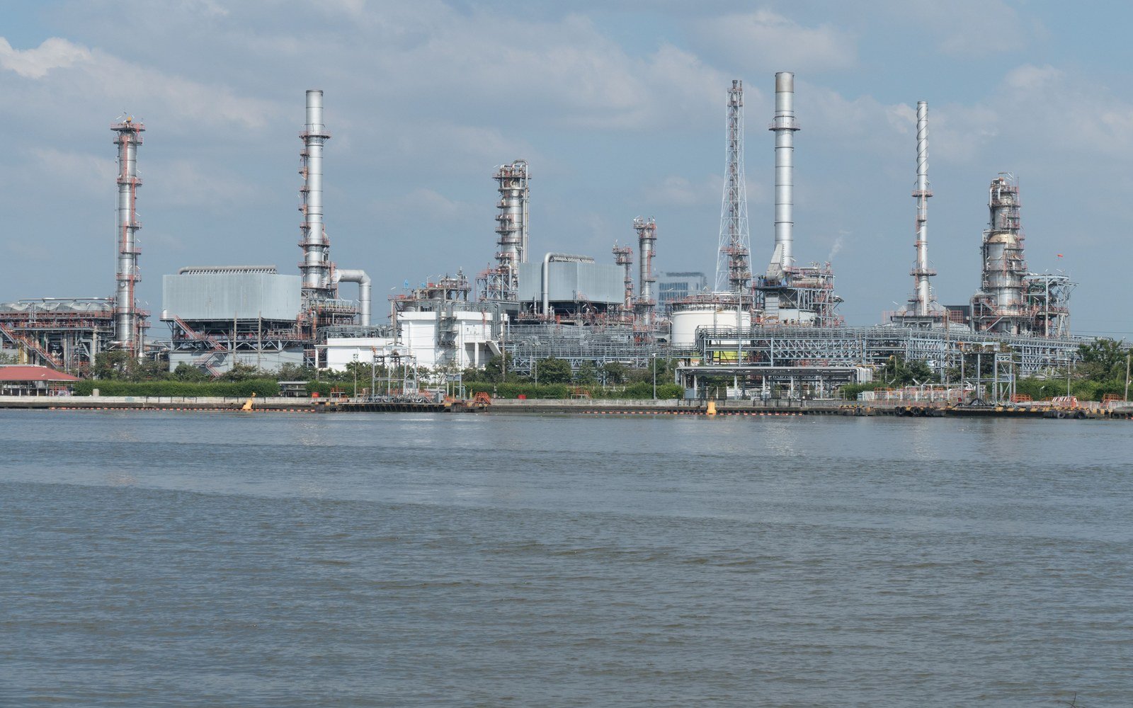 A refinery and petrochemical works seen across open water in daylight, distillation columns and stacks rising behind rows of storage tanks and pipe racks, with a quay wall along the near bank