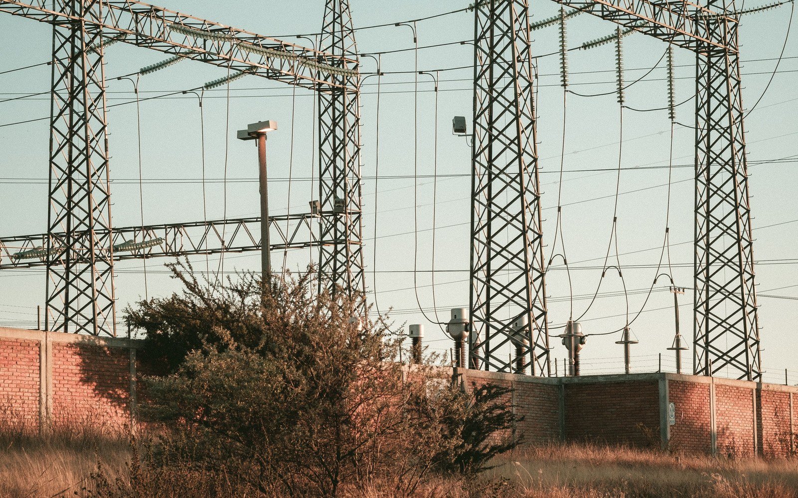 A high voltage electrical substation of steel gantries, insulators and overhead conductors standing behind scrub and a low brick wall at the edge of an open field
