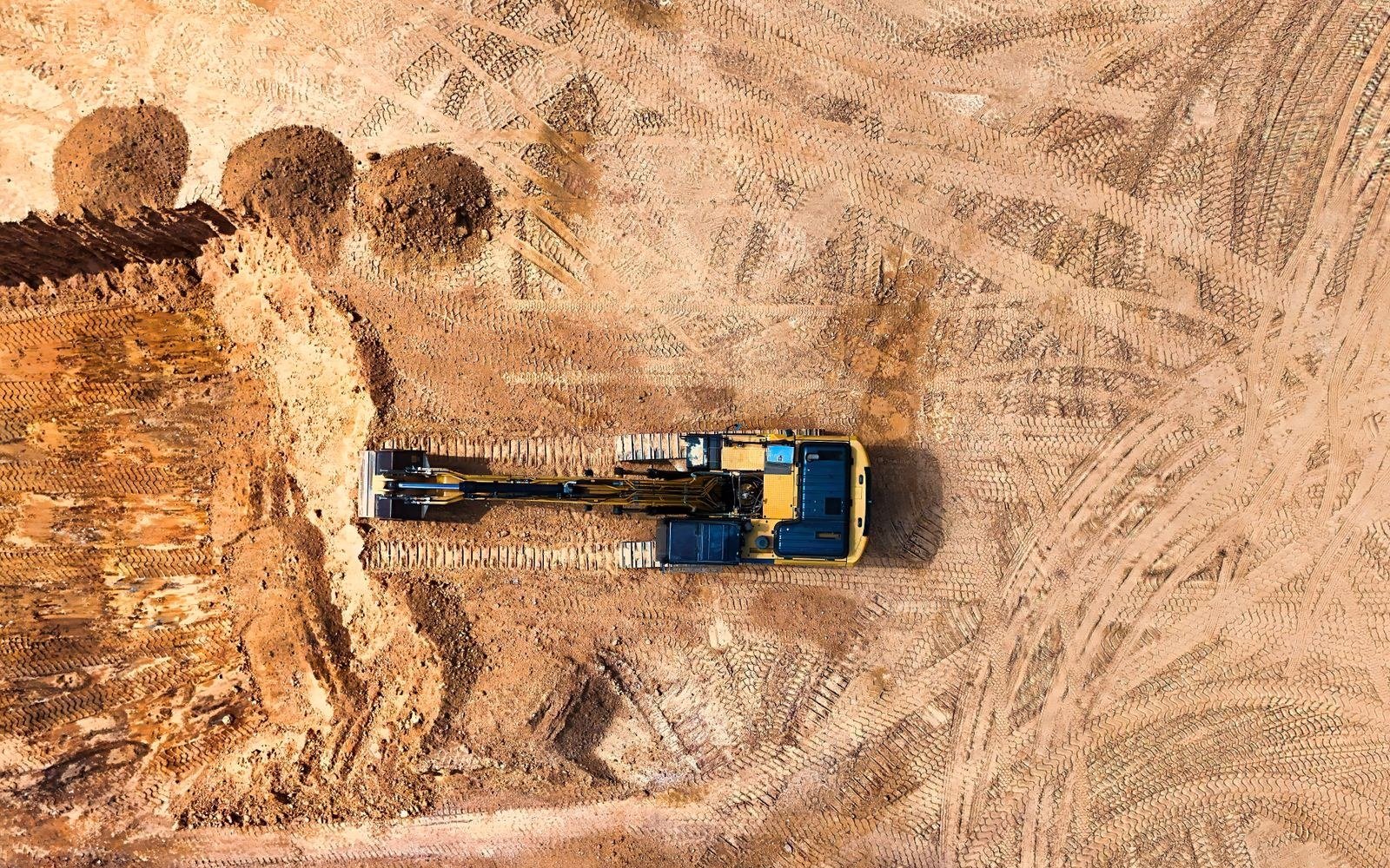 Aerial view straight down on a tracked excavator standing in a cut through dry ground, its boom laid out along the base of the cut, with tracked and wheeled plant marks crossing the ground on either side