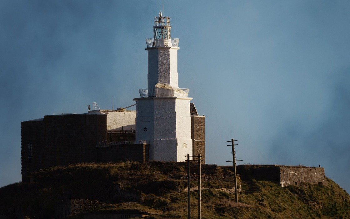 A white lighthouse on a rocky headland on the South Wales coast under a heavy grey sky, with a stone retaining wall below it