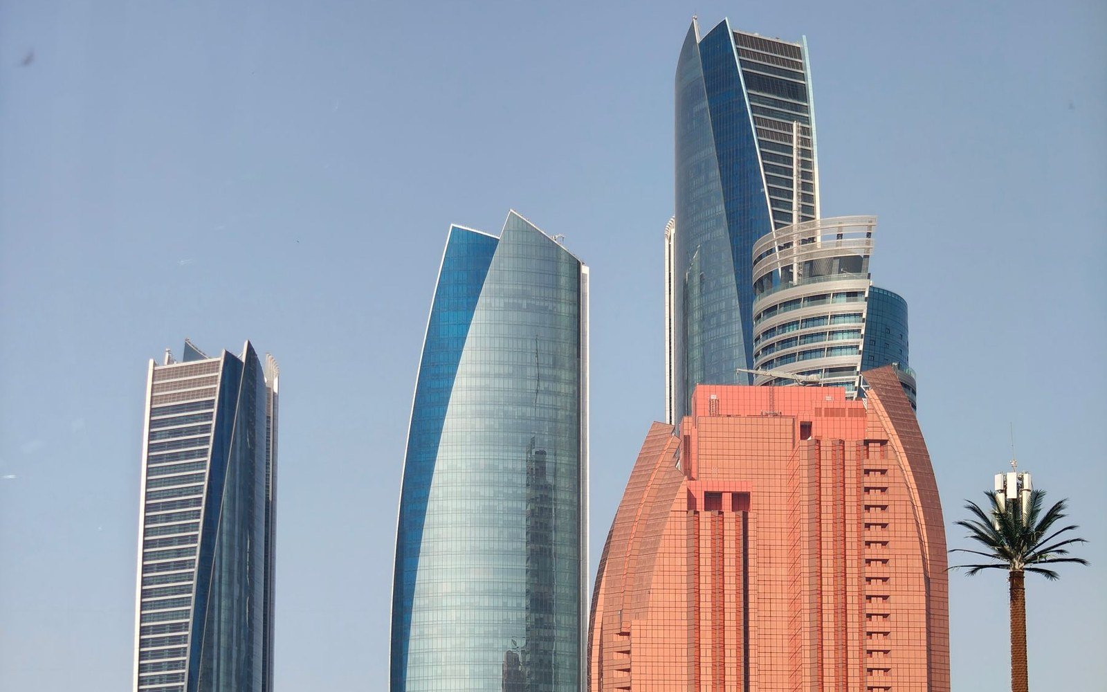 The upper storeys of the Etihad Towers and neighbouring high rise buildings in Abu Dhabi against a clear sky, with a single palm at the right