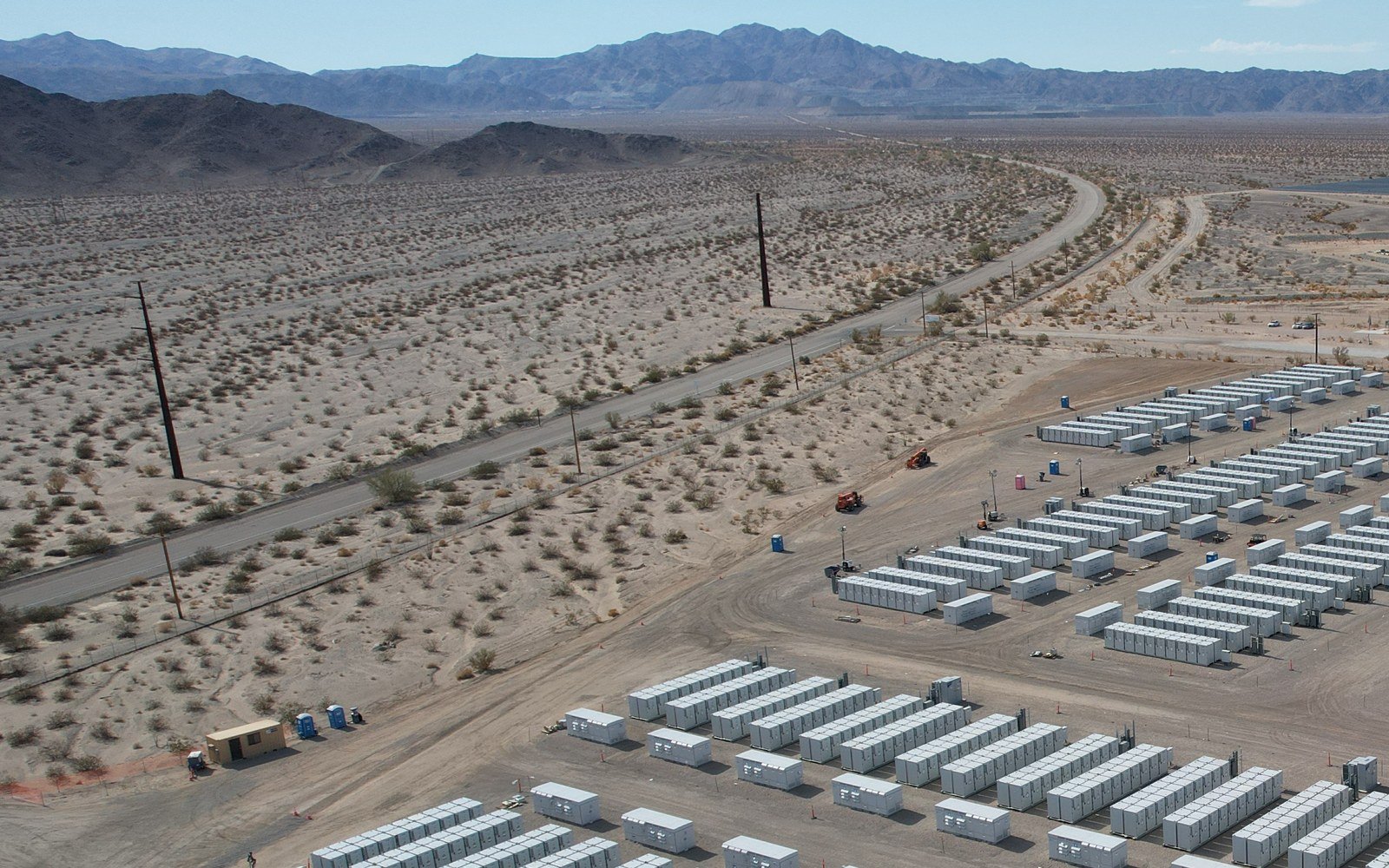 Rows of pale grey battery enclosures standing in ordered lines on a graded gravel platform in open arid country, with scrub, a two lane road and a low mountain range beyond under a clear sky