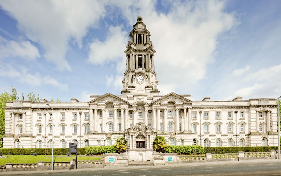 A stone civic building with a clock tower and colonnaded frontage seen from the road under a light sky