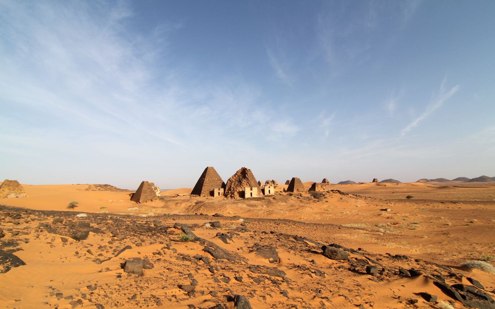 The Nubian pyramids at Meroe standing on orange desert sand under a clear sky, with rock and scrub in the foreground