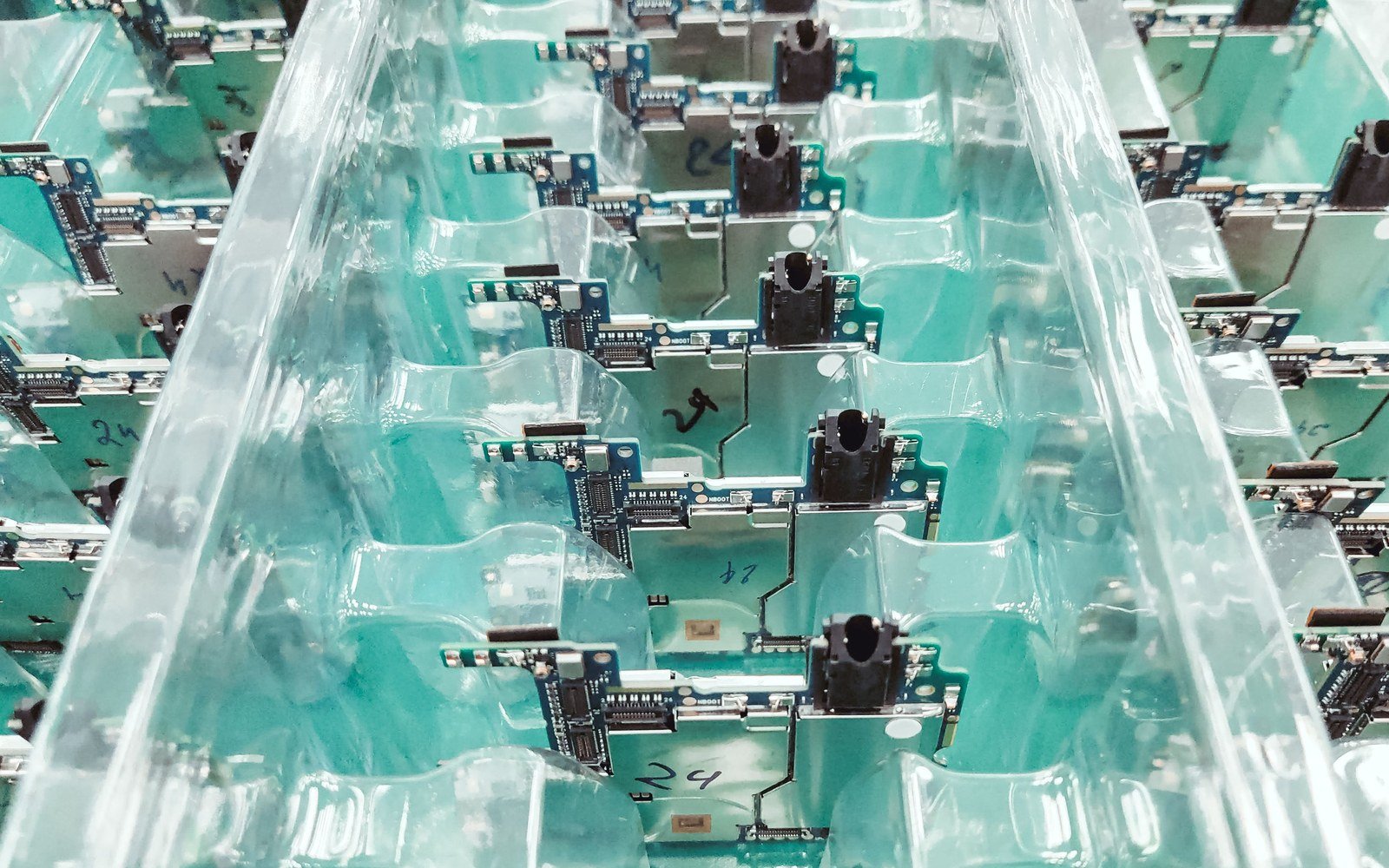 Rows of small blue circuit boards seated in a moulded clear plastic tray, each carrying a ribbon connector and a black socket, with hand written batch numbers marked on the tray between them