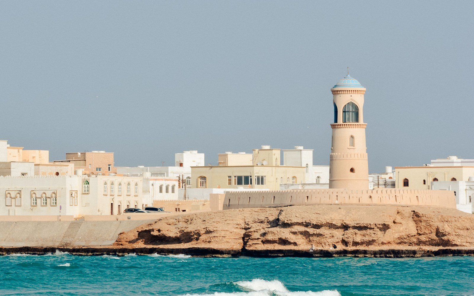 The lighthouse and fort wall on the headland at Sur in Oman, with the low white town behind and surf breaking on the rocks below