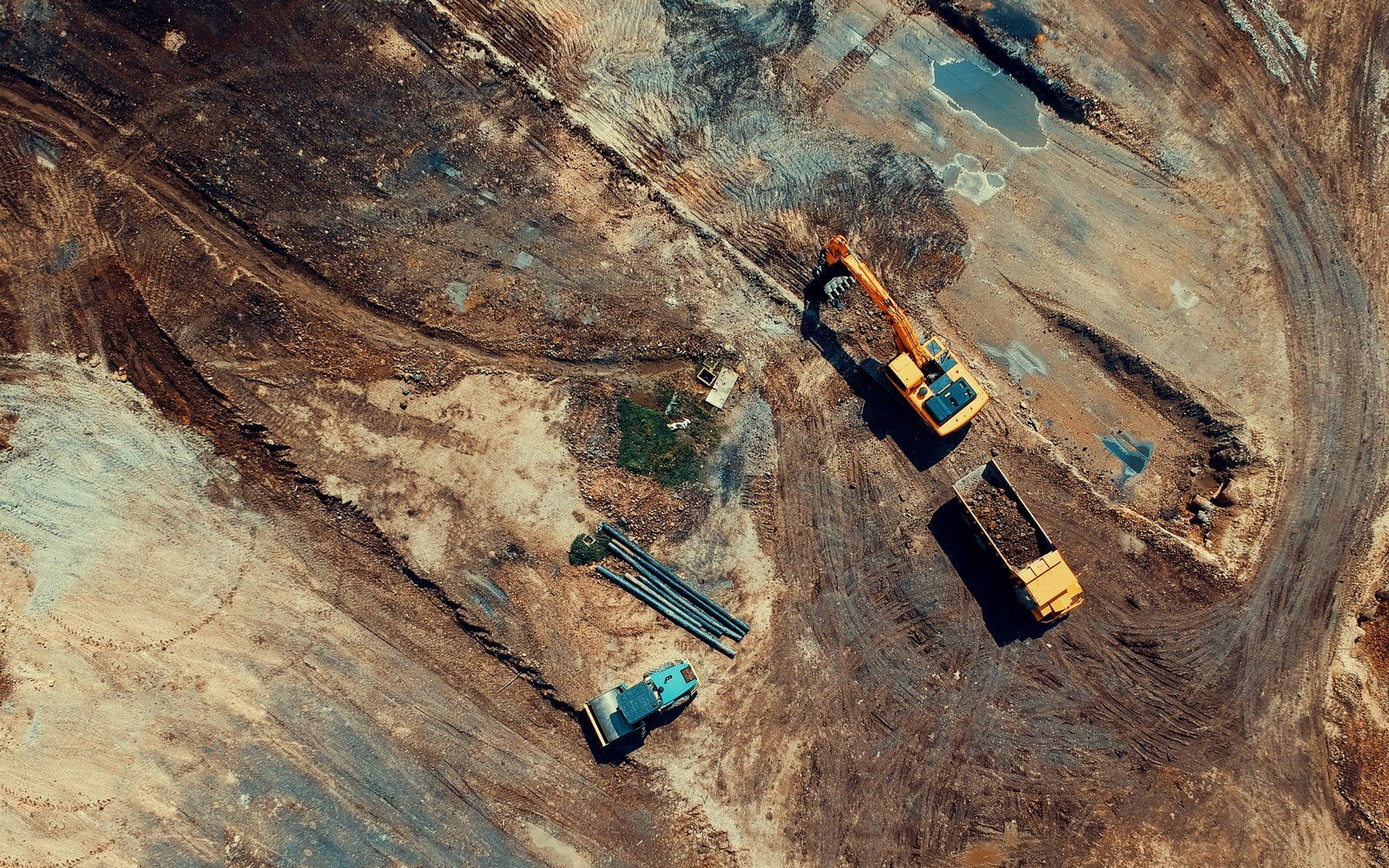 Overhead view of a stripped construction site with an excavator, a dumper and vehicle tracks across bare ground