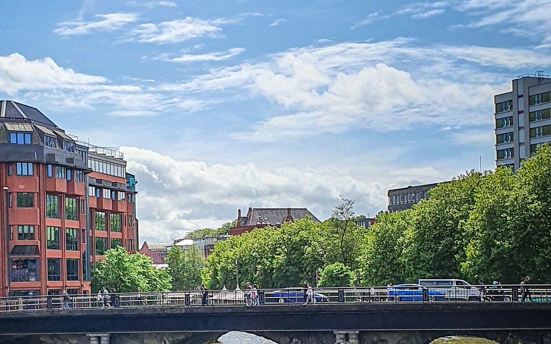 A road bridge over a river in Bristol with people walking across it, red brick riverside buildings on the left and a line of trees behind