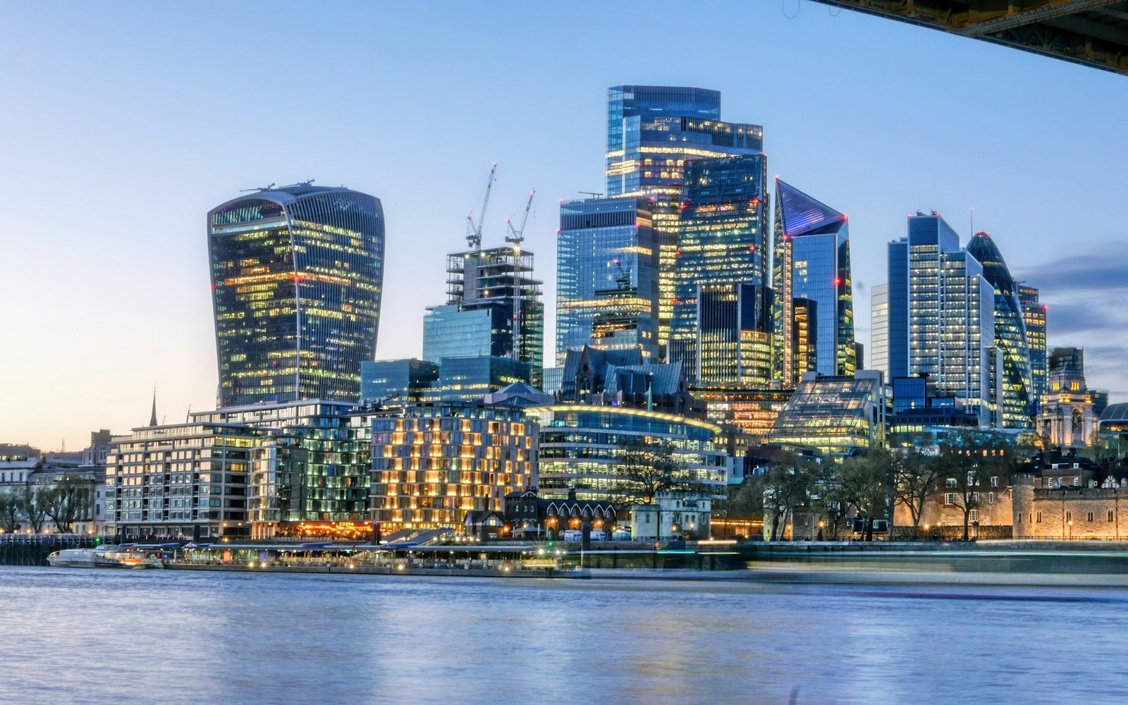 The towers of the City of london lit at dusk seen across the Thames from the south bank, with tower cranes working on two unfinished frames among the completed buildings