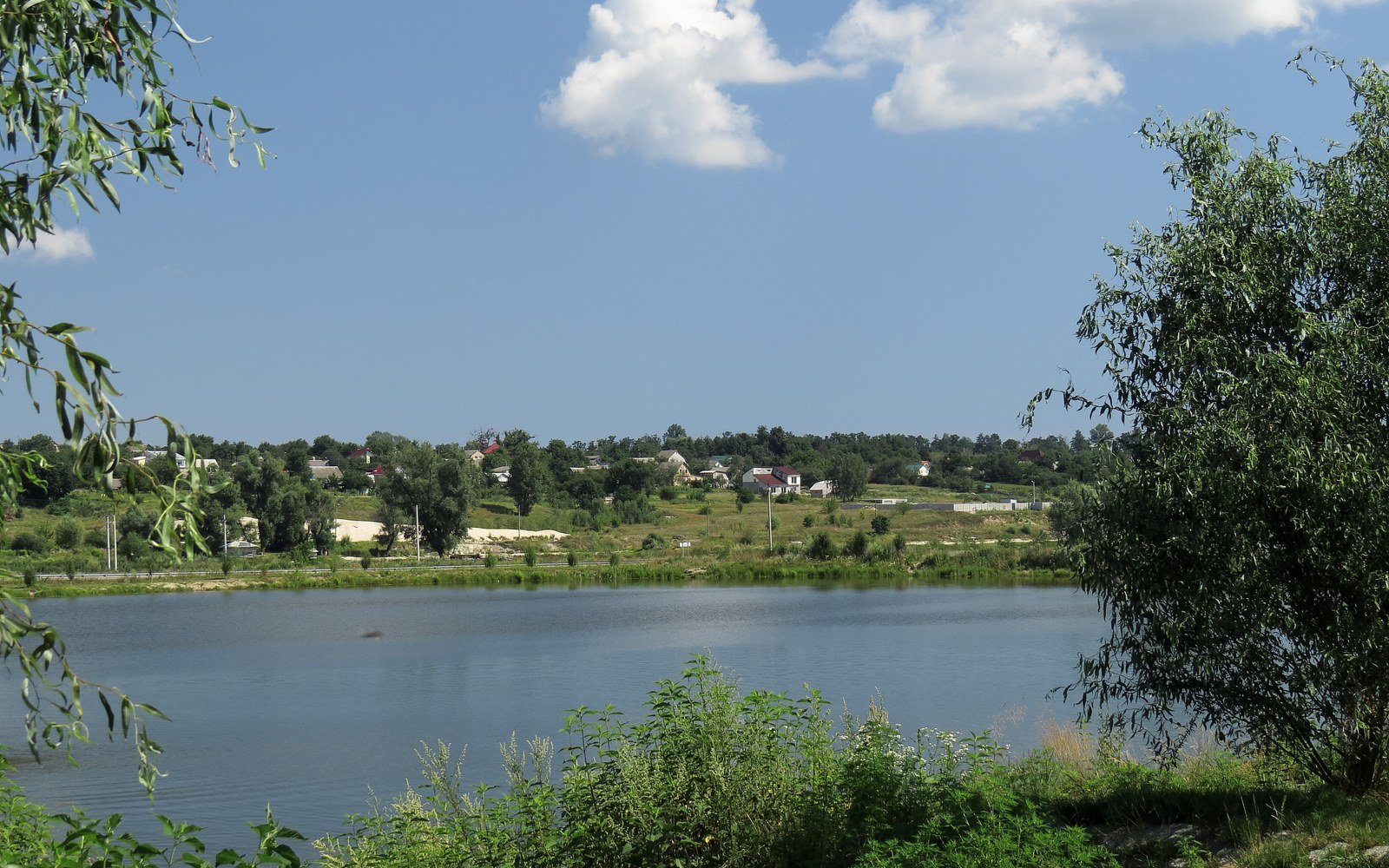 A still lake fringed by trees with low houses along the far bank in Bucha