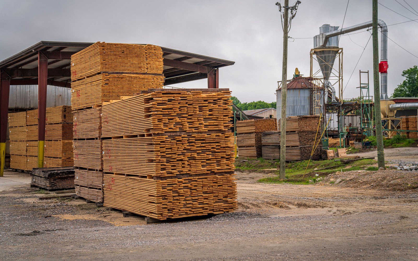 Stacks of freshly sawn boards on bearers under an open sided shed at a timber yard, with a steel silo and dust extraction cyclone behind