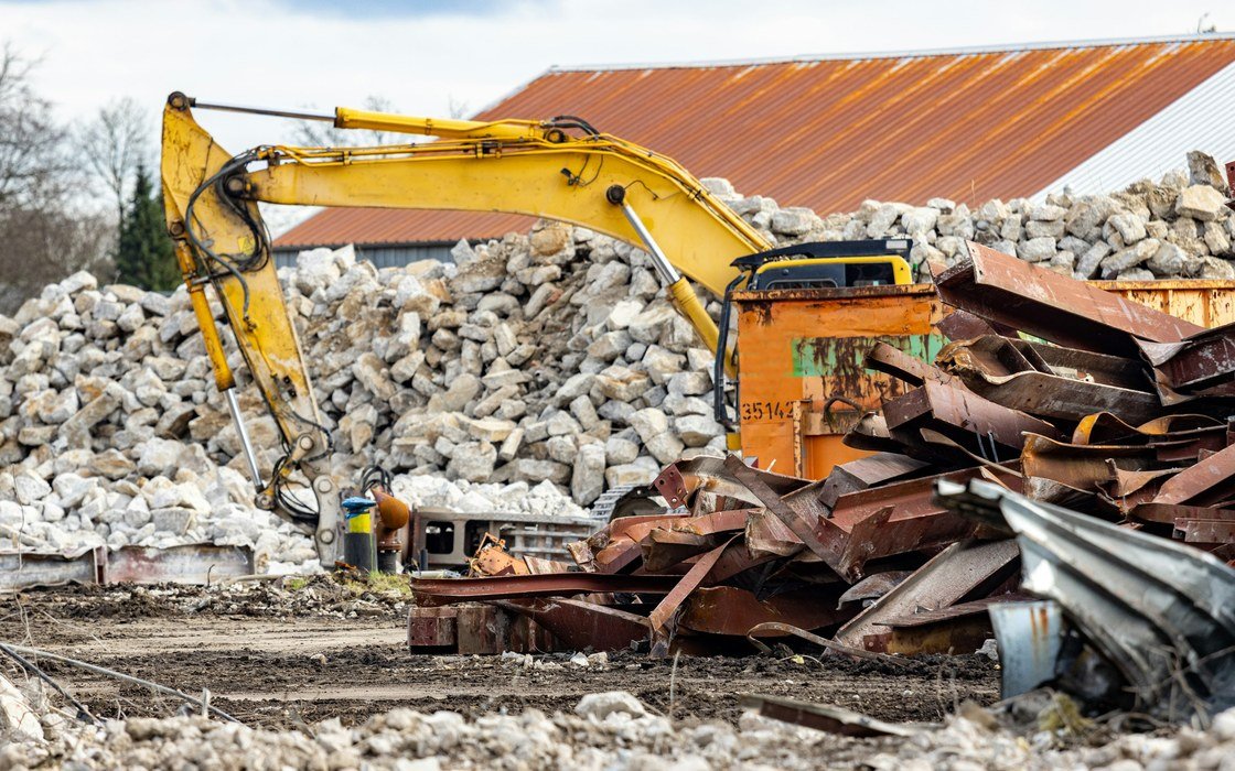 A tracked excavator standing among heaped masonry rubble and twisted sheet steel on a cleared site