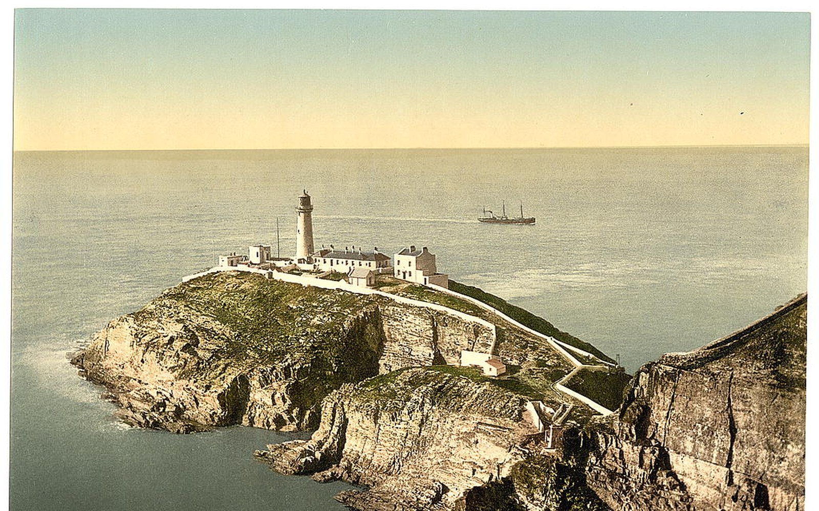 A lighthouse and its keepers' dwellings on a rocky headland above the sea, with a steamship on the water beyond