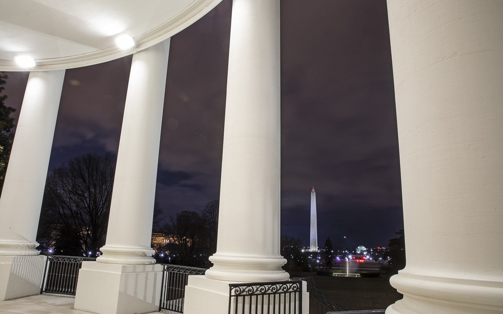 The white columns of the White House colonnade at dusk, with bare trees and the lit Washington Monument in the distance