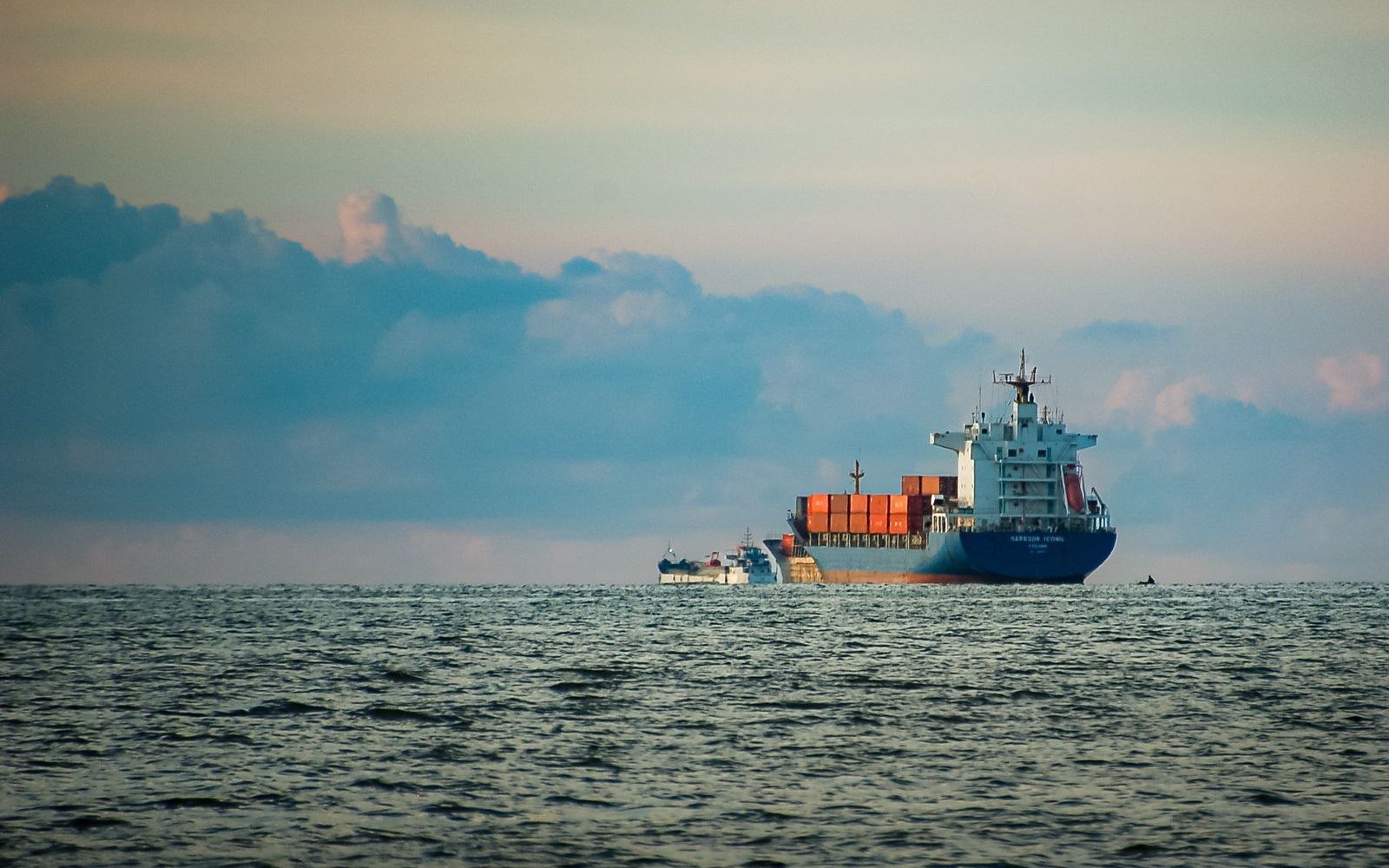 A laden container ship on the horizon in open water at dusk, with a smaller vessel astern of it and cloud banked along the skyline